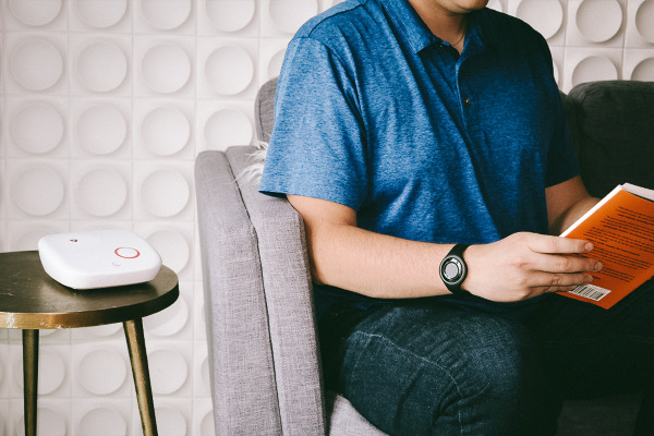 Person reading book wearing a black help button bracelet next to Home 2.0 on side table