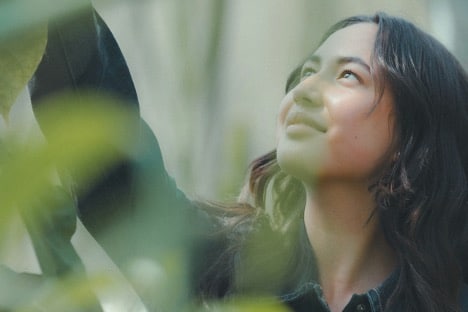 A woman gazes upward, admiring the branches and leaves of a tall tree, exuding self-esteem