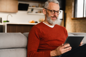 Man in red sweater sitting on a couch and holding a tablet in his hands.